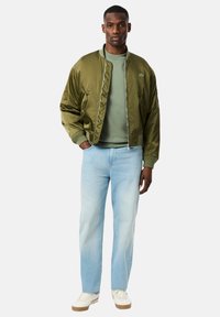 Chaqueta bomber de satén verde oliva, camiseta de cuello redondo verde claro, vaqueros azul claro, zapatillas blancas. La chaqueta tiene puños acanalados y un detalle de logotipo.