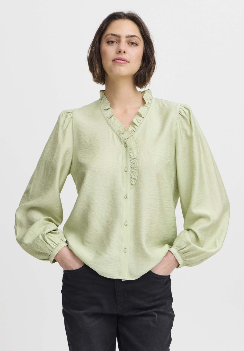 Light green blouse featuring a ruffled neckline, long puff sleeves, and button closure. The material has a soft, textured finish.