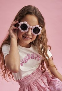 Roze bloemvormige zonnebril met donkere lenzen, gedragen door een meisje in een witte shirt met "SUNSHINE" in roze glitters, en een roze gebloemde rok.