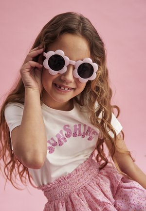 Rosa blütenförmige Sonnenbrille mit dunklen Gläsern, getragen von einem Mädchen in einem weißen Shirt mit "SUNSHINE" in rosa Glitzer und einem rosa gemusterten Rock.