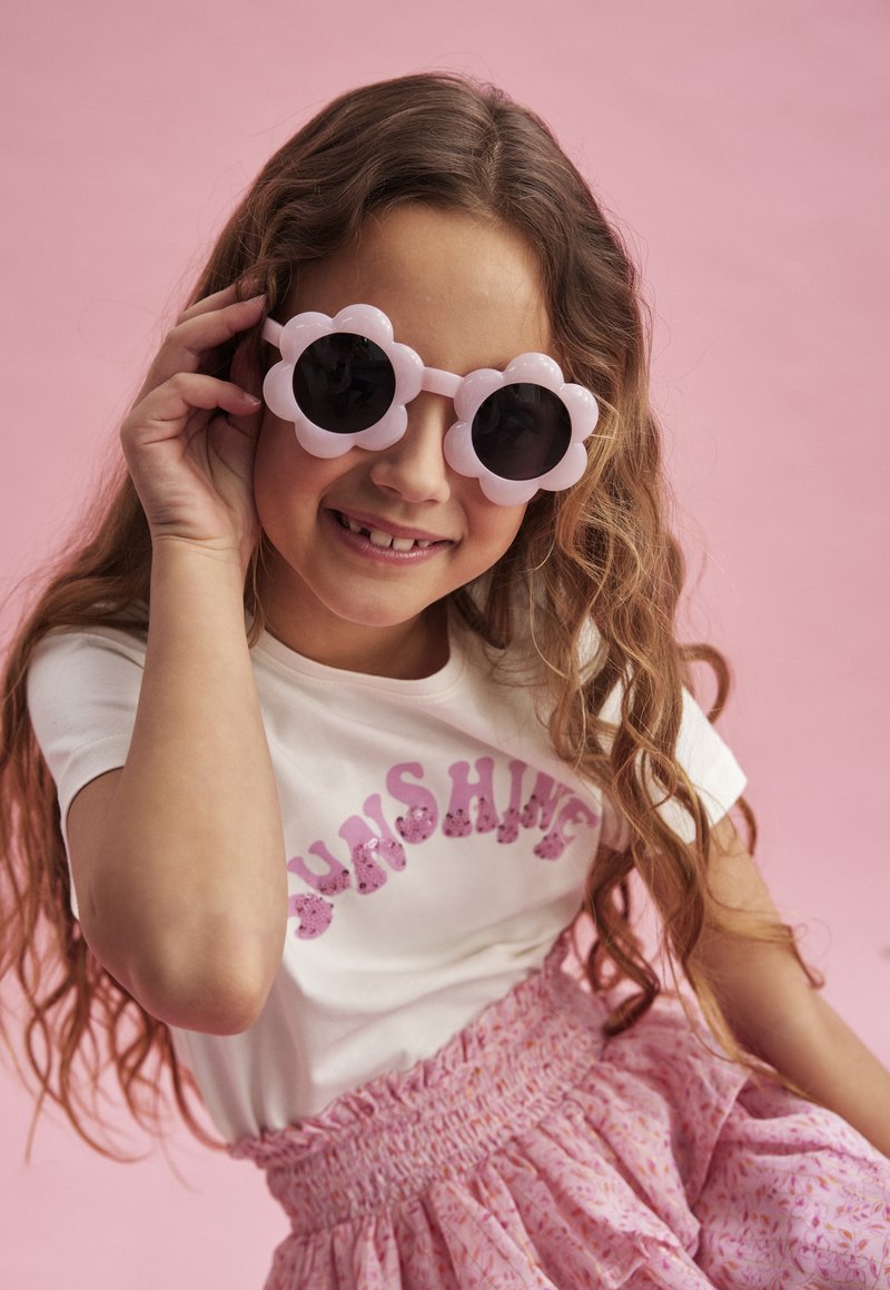 Roze bloemvormige zonnebril met donkere lenzen, gedragen door een meisje in een witte shirt met "SUNSHINE" in roze glitters, en een roze gebloemde rok.