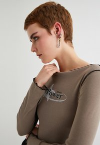 Pitkähiha, istuva toppi taupen värissä mustilla yksityiskohdilla, jossa on harmaa tähden graafinen kuvio ja sana "Voima". Asusteina ovat korvakorut ja rannekoru.