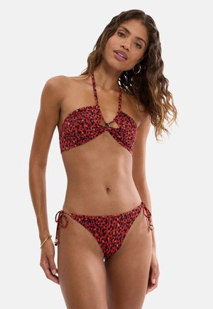 Kobieta o falowanych włosach, ubrane w czerwono-czarny bikini na szyję w panterkę z wiązaniami po bokach, stojąca na jednolitym jasnym tle.