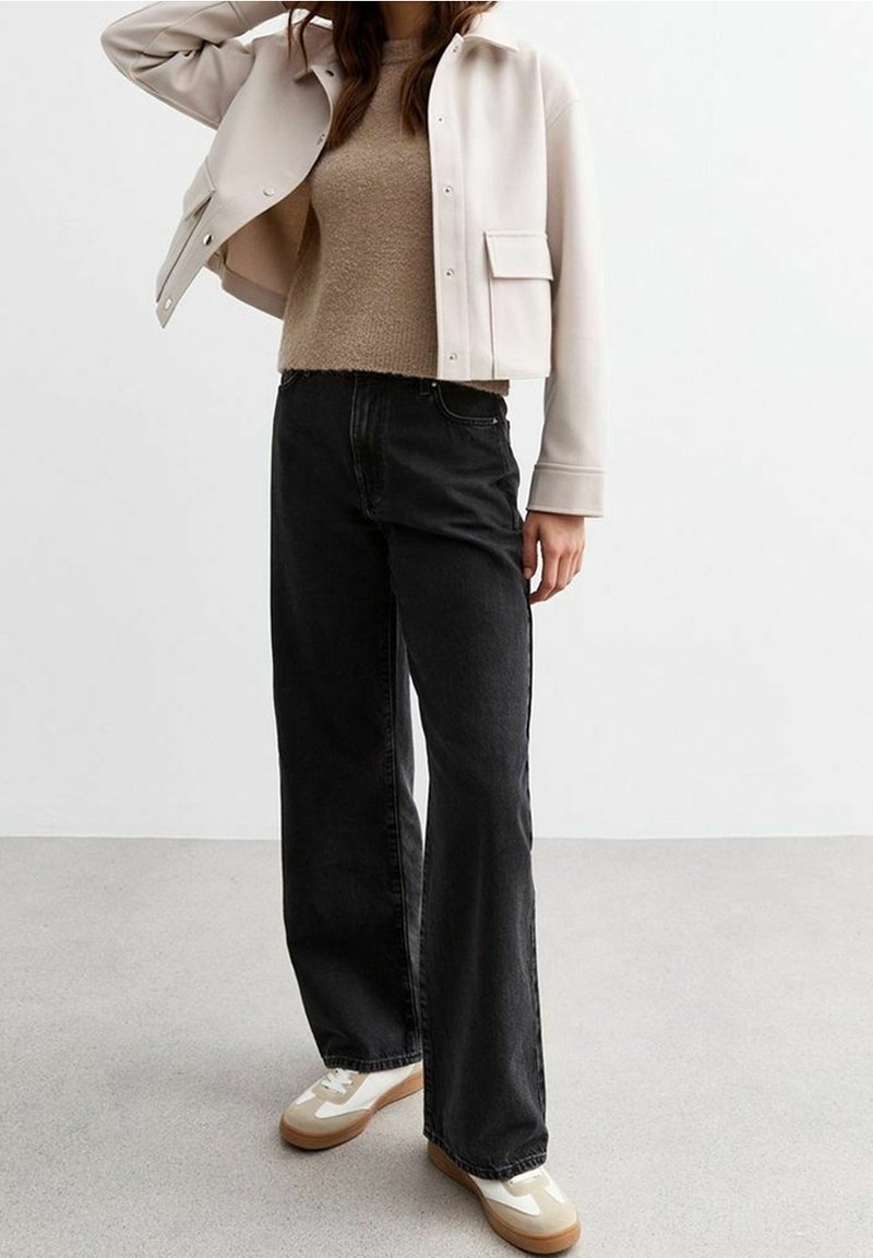 Femme portant une veste crème, un pull beige, un jean noir à jambes larges et des baskets blanches, debout devant un fond uni.