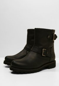 Bottes chevilles en cuir noir avec une semelle plate, dotées d'une lanière latérale et d'un détail de boucle, texture lisse et construction robuste.