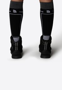 Chaussettes noires montantes avec un bord gris, présentant des détails en logo blanc et des rayures, portées avec des bottes de randonnée noires, sur un fond neutre.
