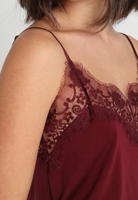 Camisola bordeaux con delicata rifinitura in pizzo, caratterizzata da un bordo ondulato. Il materiale sembra liscio con sottili spalline regolabili.