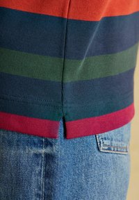 Detailný záber na lem pruhovanej pletenej košele s červenými, zelenými a námornícky modrými pruhmi, ktorý sa nachádza nad vreckom modrých džínsov.