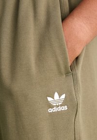 Ruka odpočívající uvnitř kapsy olivově zelených kalhot Adidas s bílým logem trefoil.