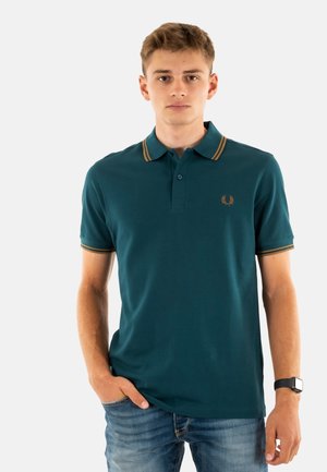 Polo en verde azul profundo con cuello clásico, tapeta de dos botones y rayas horizontales en dorado. Presenta un pequeño logo en el pecho.