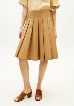 Frau trägt einen hellbraunen, plissierten Rock knielang, ein beiges kurzärmeliges Hemd und braune, offene Blockabsatz-Mules und steht vor einem einfarbigen Hintergrund.