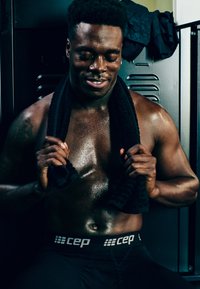 Muscular individual wearing a black CEP waistband, holding a black towel. Skin appears shiny and wet, with a dark background and lockers.