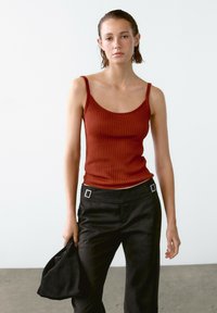 Gestreiftes, rostfarbenes Tanktop mit dünnen Trägern, kombiniert mit hochtaillierten, schwarzen Hosen, die rechteckige silberne Hardware-Akzente aufweisen.