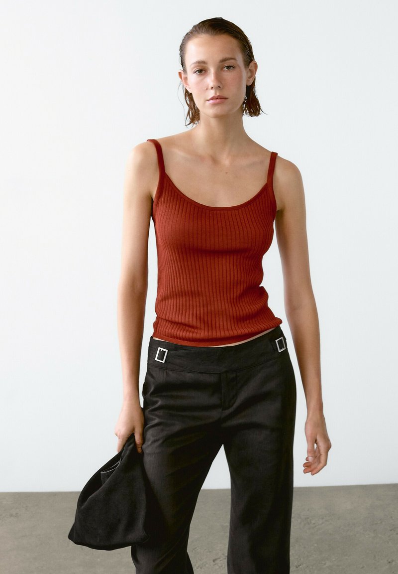 Gestreiftes, rostfarbenes Tanktop mit dünnen Trägern, kombiniert mit hochtaillierten, schwarzen Hosen, die rechteckige silberne Hardware-Akzente aufweisen.