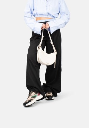 Borsa a tracolla bianca con design curvo, tracolla accentuata e texture liscia. Indossata con pantaloni neri larghi e sneaker multicolori.