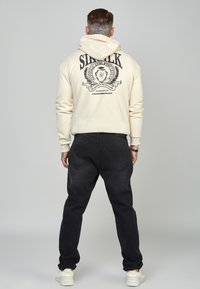 Hombre con el cuello tatuado, vistiendo una sudadera crema con texto impreso y emblema en la parte trasera, jeans negros y zapatillas blancas, de pie mirando hacia atrás.