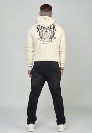 Hombre con el cuello tatuado, vistiendo una sudadera crema con texto impreso y emblema en la parte trasera, jeans negros y zapatillas blancas, de pie mirando hacia atrás.