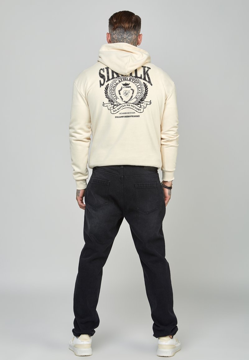 Hombre con el cuello tatuado, vistiendo una sudadera crema con texto impreso y emblema en la parte trasera, jeans negros y zapatillas blancas, de pie mirando hacia atrás.