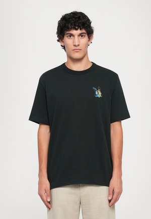 Joven con cabello rizado que lleva una camiseta negra con un pequeño diseño de un conejo en arte de píxeles de colores en el lado izquierdo del pecho, pantalones beige.