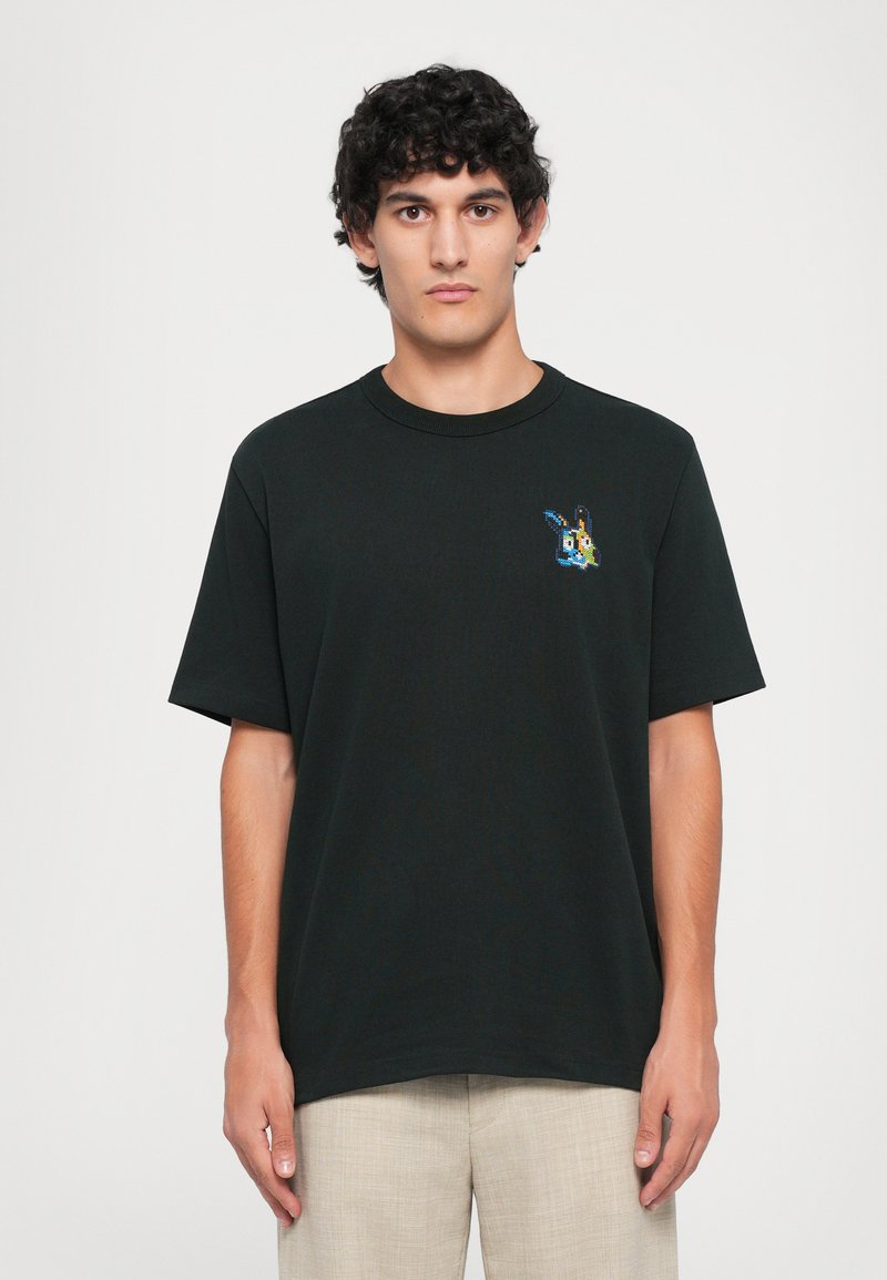 Tânăr cu părul creț, purtând un tricou negru cu un mic design de iepure pixelat colorat în partea stângă a pieptului, pantaloni bej.