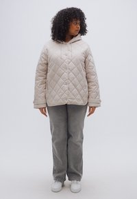 Bež prešita puhovka z ruto, z vzorcem v obliki diamantov in sprednjimi pritisnimi gumbi, kombinirana s sivimi kordurovnimi hlačami in belimi supergami.