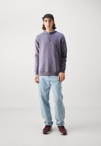 Fialová mikina s kapucňou a prednou vreckom, v kombinácii s svetlomodrými džínsami a tmavočervenými teniskami. Na pozadí bez vzoru.