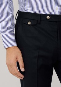 Crne hlače teksture sa dualnim dugmadi na struku, uparene s plavom i bijelom prugastom košuljom, koja ima standardni dizajn manžeta.