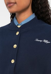 Námornícky modrá mikina s okrúhlym golierom, zlatými gombíkmi a vyšitým logom "Tommy Hilfiger" v bielej farbe. Má textúrovanú tkaninu.
