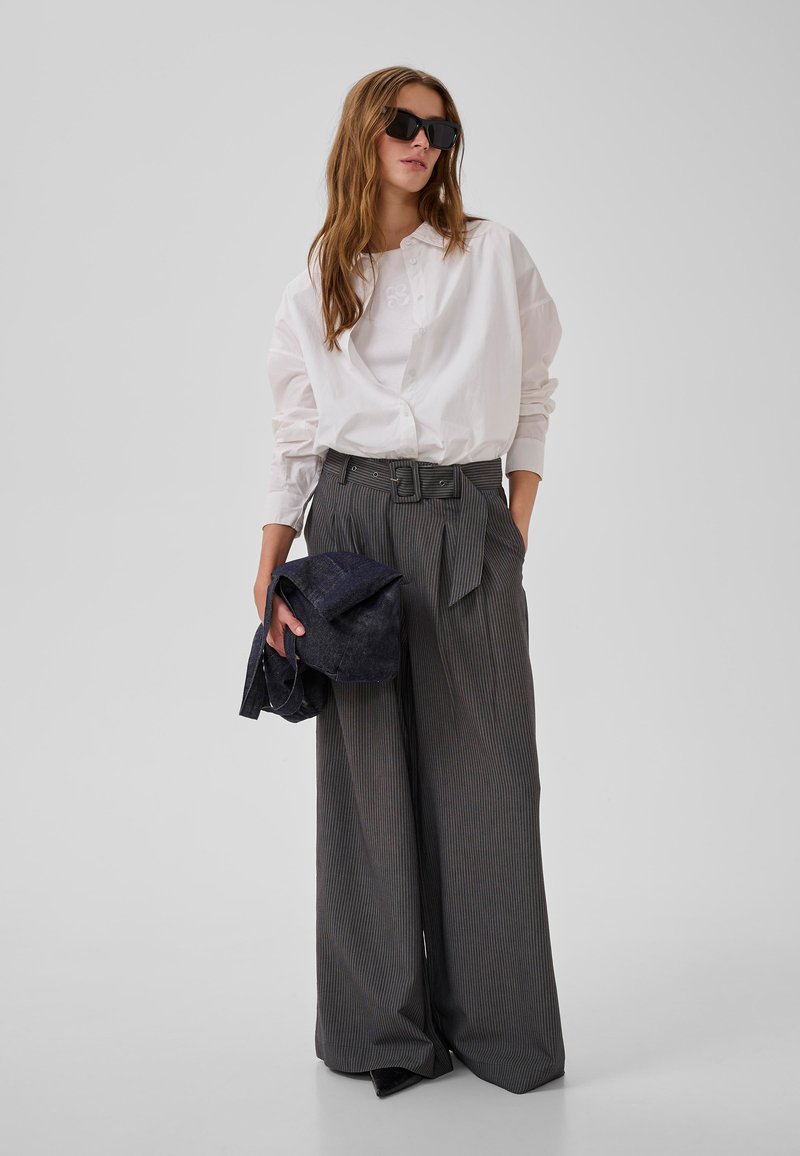 Ženska v preveliki beli srajci, širokih hlačah z ozkimi črtami, s črnimi sončnimi očali, drži zloženo temno jakno na enobarvnem ozadju.