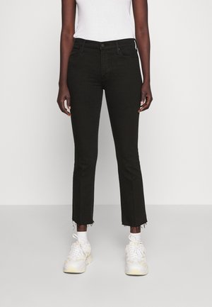 Person iført sorte cropped jeans med flossede kanter, en hvid tætsiddende top, hvide sokker og hvide sneakers, der står mod en ensfarvet baggrund.