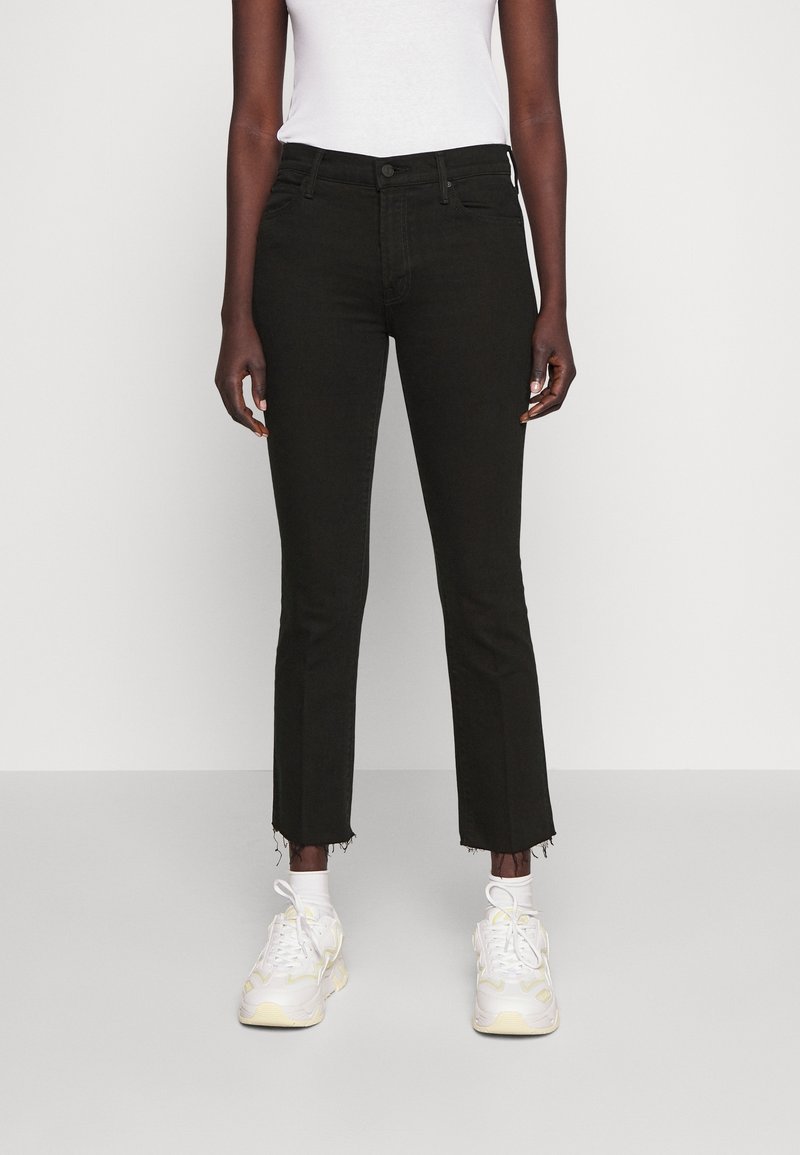 Personne portant un jean noir court avec des ourlets effilochés, un haut blanc ajusté, des chaussettes blanches et des baskets blanches, debout devant un fond uni.