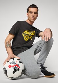 Mand iført sort BVB fodboldtrøje og grå sweatpants knæler med hånden på en sort og hvid fodbold fra Puma.