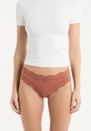 Slip couleur rouille orné de dentelle et d'un petit nœud ; associé à un haut blanc moulant à manches courtes. Le tissu semble doux et extensible.