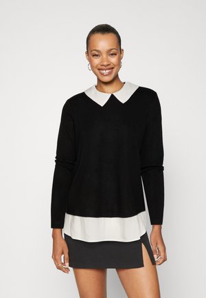 Un pull noir à manches longues avec un col blanc, superposé sur l'ourlet d'une chemise blanche. Le mannequin porte une mini-jupe noire, mettant en valeur une fente sur le côté.