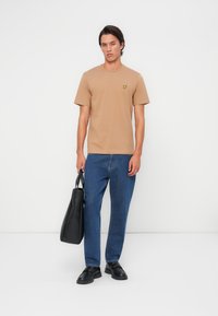T-shirt beige à manches courtes avec un petit logo jaune sur la poitrine, associé à un pantalon en denim bleu et des chaussures noires, portant un sac noir.