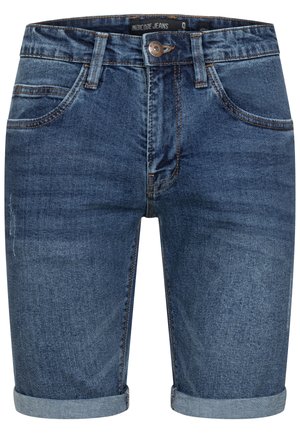 Mand iført opkørte mørkeblå denimshorts og hvide sneakers, stående med ryggen mod kameraet mod en hvid baggrund.