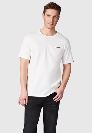 Man draagt een eenvoudige witte T-shirt met korte mouwen en een klein geborduurd vliegtuigje op de borst, gecombineerd met een zwarte spijkerbroek, staand tegen een effen achtergrond.
