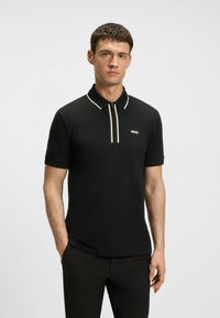 Schwarzes Polohemd mit Reißverschlusskragen, mit weißen und goldenen Akzenten sowie einem kleinen "BOSS"-Logo auf der linken Brust. Kurzärmelig.
