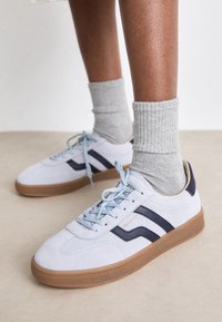 Pieds portant des baskets gris clair avec des semelles en gomme et des bandes sombres en forme de vague, associées à des chaussettes côtelées gris clair retroussées sur un sol neutre.