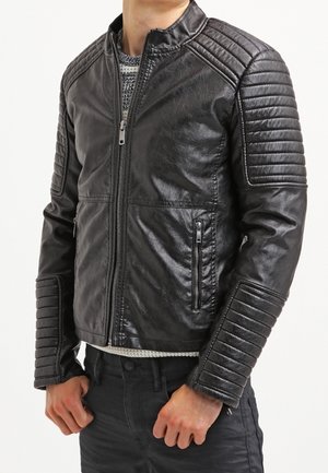 Mężczyzna noszący czarną pikowaną skórzaną kurtkę z kieszeniami na suwak na szarym swetrze i czarnych spodniach, ręce spoczywające blisko talii.