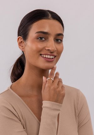 Silberring mit drei weißen Perlen, am rechten Handgelenk getragen. Das Model hat einen glatten, hellen Teint und lange, dunkle Haare. Sie trägt ein beiges, geripptes Oberteil.
