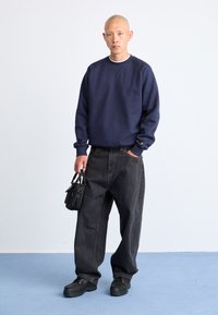 Hombre calvo de pie en un suelo azul, vistiendo un sudadero azul marino, jeans negros holgados, zapatos negros, sosteniendo un pequeño bolso negro con una mano en el bolsillo.