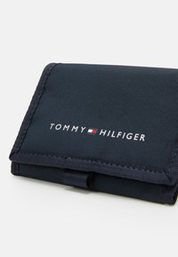Granatowy portfel z teksturowanej tkaniny z zapięciem na klapkę, z logo "TOMMY HILFIGER" w białych i czerwonych akcentach.