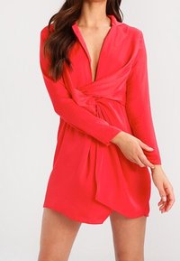 Robe rouge à manches longues avec un décolleté en V profond et un détail torsadé à la taille. Tissu léger avec une texture douce, longueur au-dessus du genou.