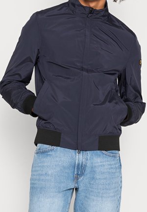 Personne portant une veste bomber bleu marine zippée, avec poignets et bas côtelés, associée à un jean bleu clair, mains dans les poches de la veste.