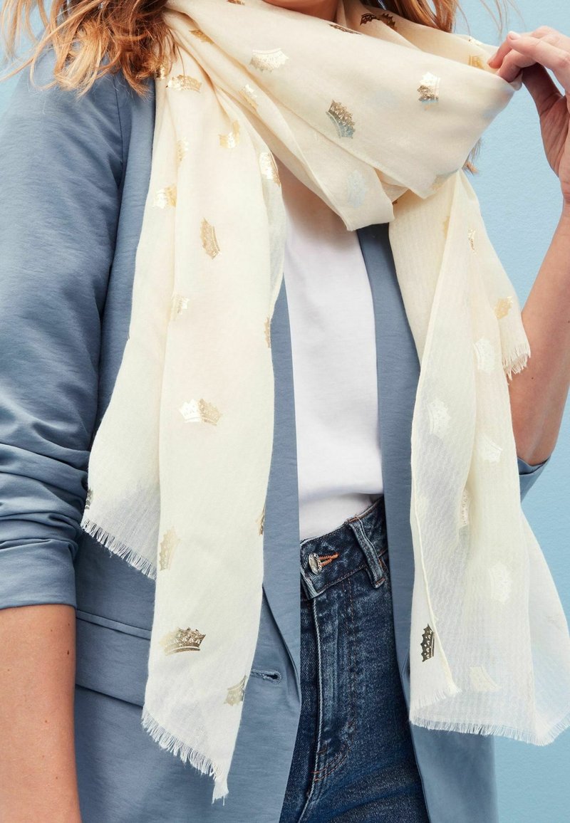 Écharpe crème claire avec motifs de couronnes dorées drapée autour du cou sur une chemise blanche et un blazer bleu clair, associée à un jean bleu.