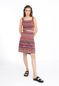 Twee-delig outfit met een kleurrijk patronen crop top en een bijpassende rok, beide in tinten roze, blauw en groen. Platte zwarte sandalen.