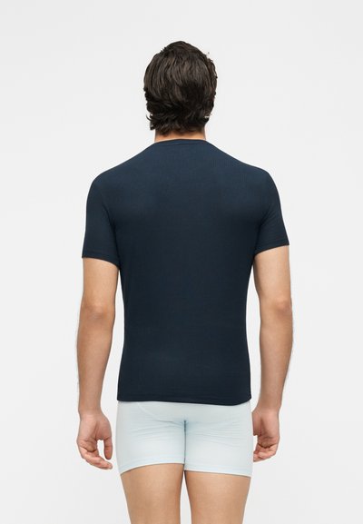 Άνδρας με καστανά μαλλιά που φοράει στενό σκουρομπλε κοντομάνικο πουκάμισο και ανοιχτό μπλε σορτς, στέκεται με την πλάτη σε απλό φόντο.