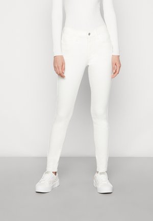 Femme portant un jean blanc slim, un haut blanc côtelé à manches longues et des baskets compensées blanches, debout devant un fond uni.