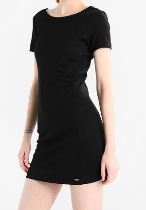 Kapeamalli musta lyhythihainen mekko pyöreällä kaula-aukolla, yllään henkilö, jonka kädet ovat rentoina sivuilla, yksivärinen valkoinen tausta.